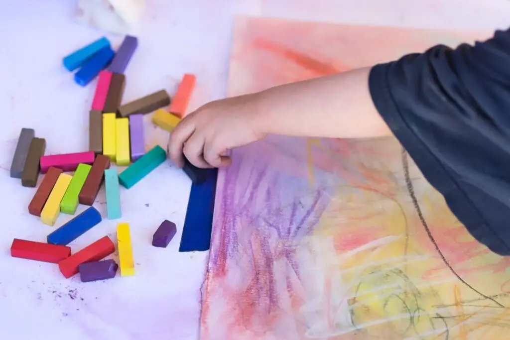 An artist hand picking up pastels to draw and different color pastels lying on the floor near a pastel drawing