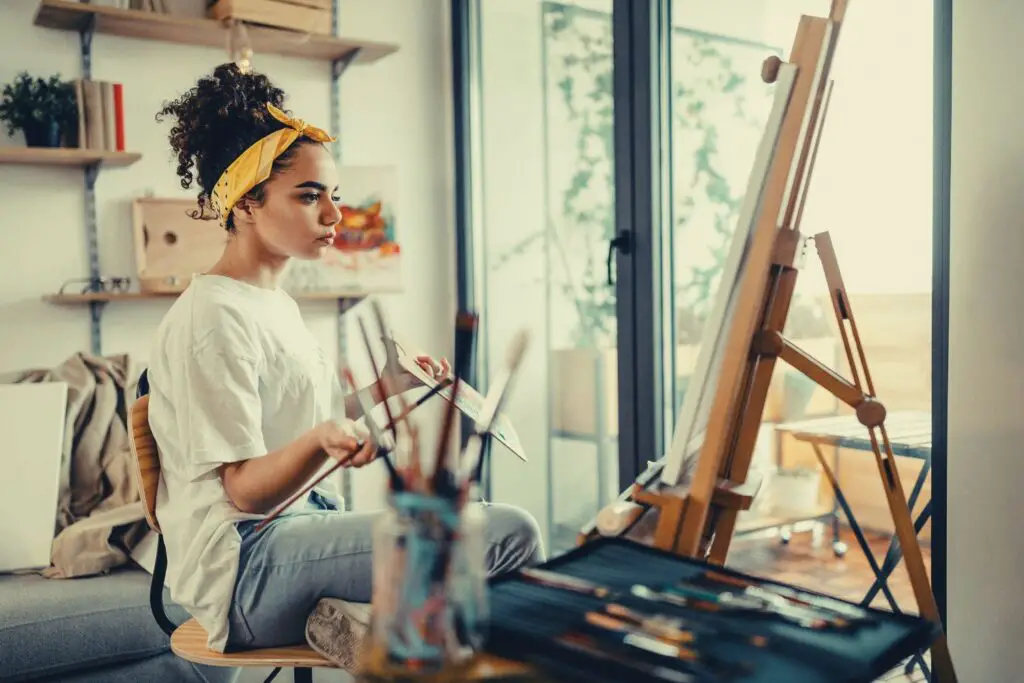 A lady artists painting on her easel sitting with brush and pens around her