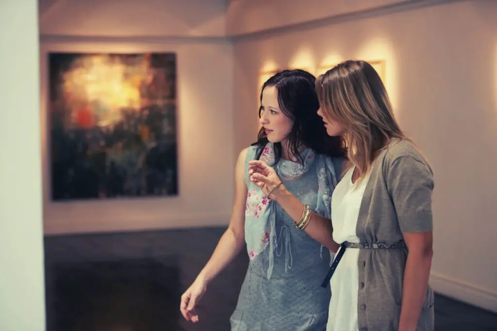 Two ladies looking at an artwork and are in discussion, and in the background there is a huge painting