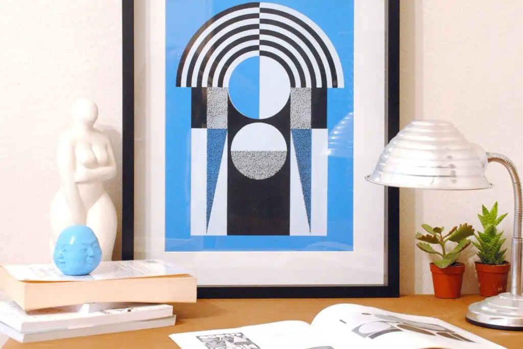 A framed Art print containing simple design on a table with books and table lamp in front.